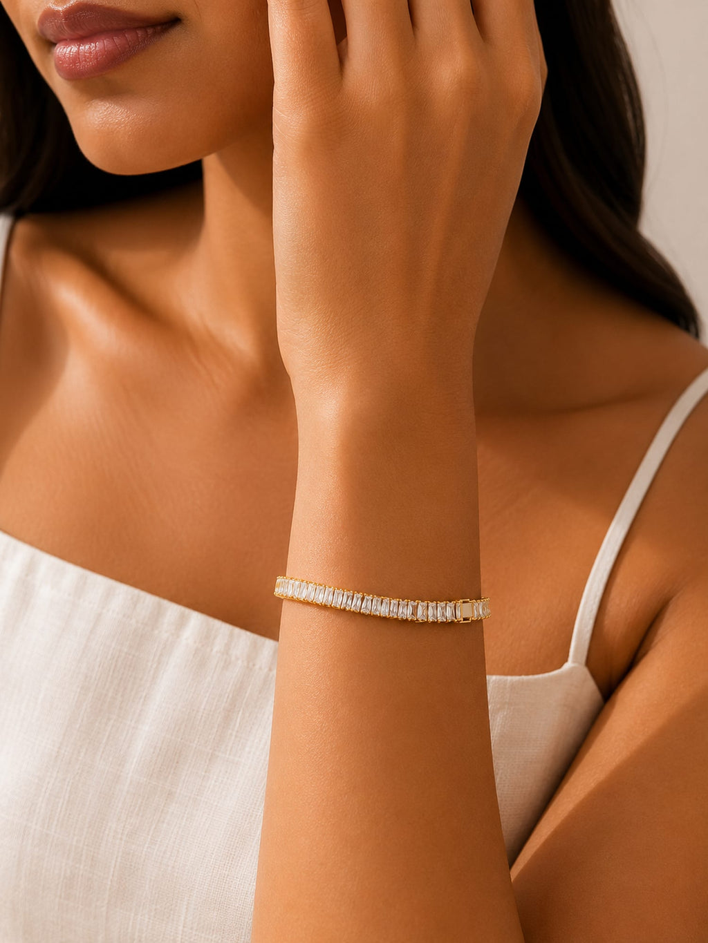 A close-up of a woman wearing a delicate gold tennis bracelet with clear stones on her wrist, paired with a white spaghetti strap top.