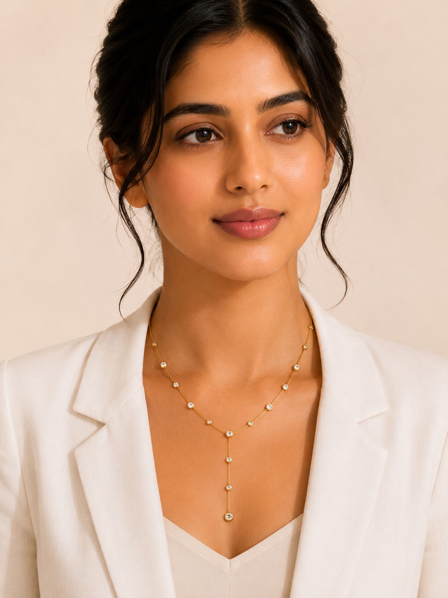 A woman wearing a delicate gold Y-necklace with small round pearl-like beads and a central drop, styled with a white blazer against a soft beige background.