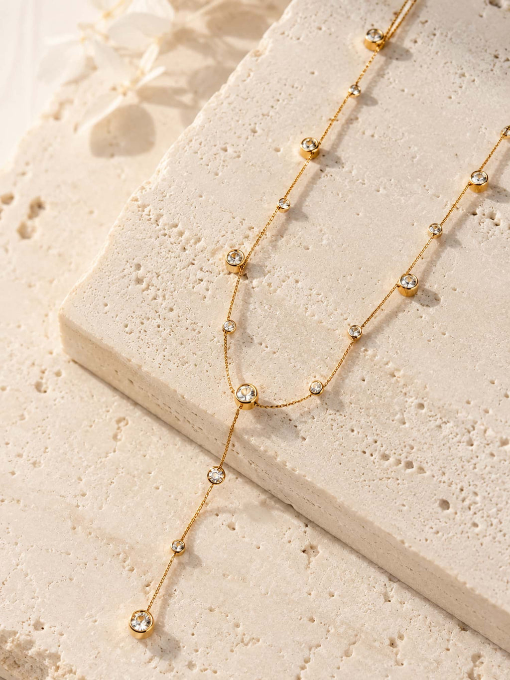 A delicate gold Y-necklace with small clear cz stones, styled on a textured beige stone slab.