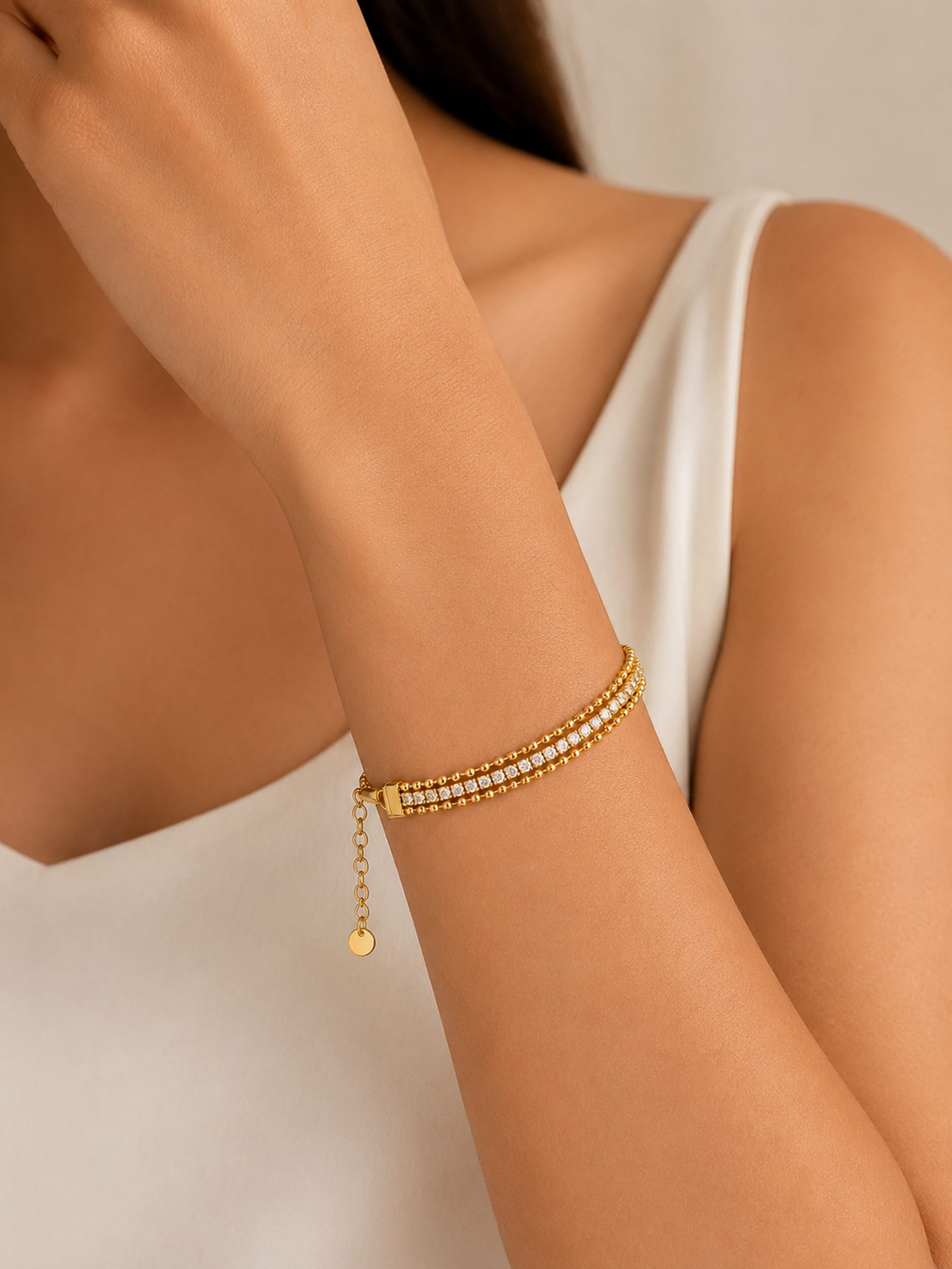 A close-up of a model's wrist wearing a delicate gold chain bracelet with two rows of small clear stones and a dangling gold charm, paired with a white sleeveless top.