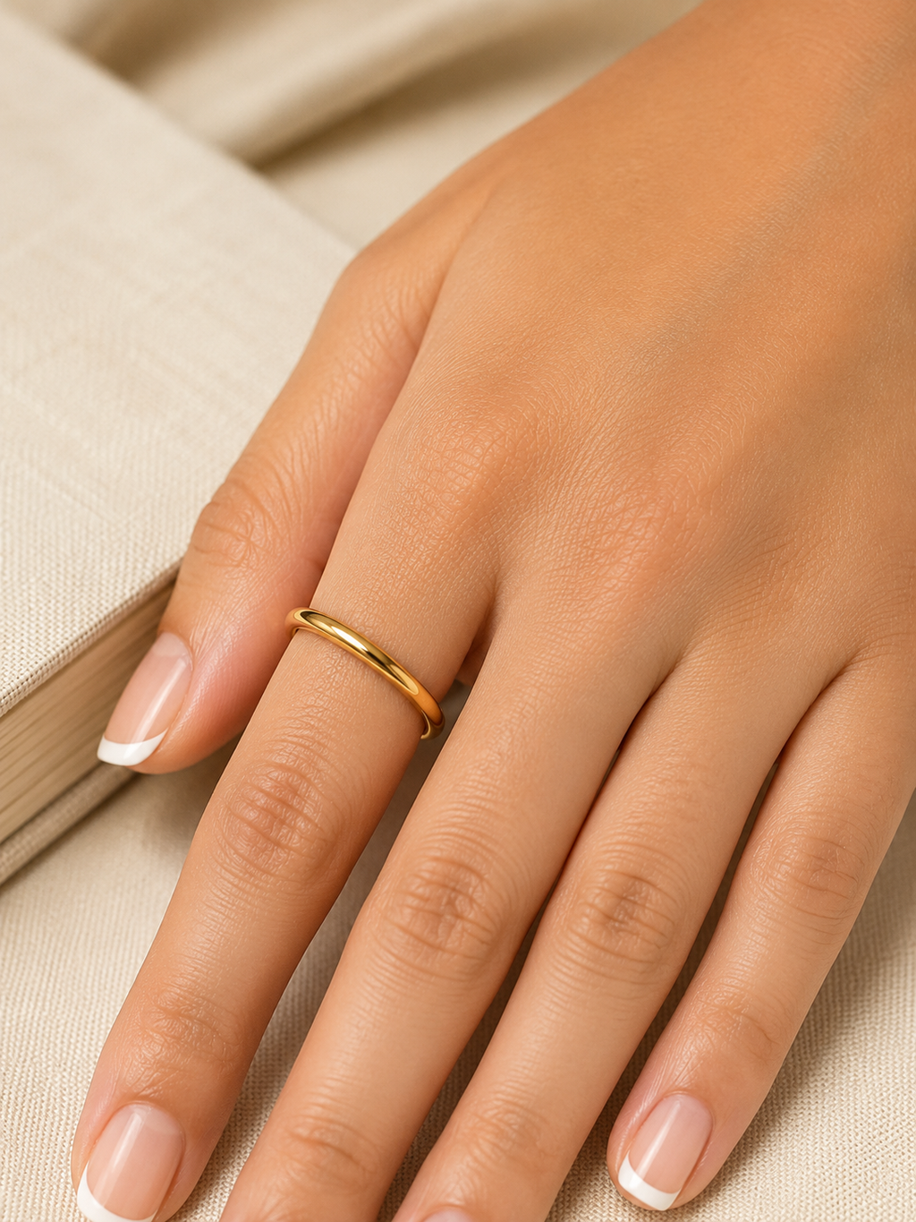A close-up of a hand wearing a thin, polished gold band ring, resting on a neutral beige textured surface.