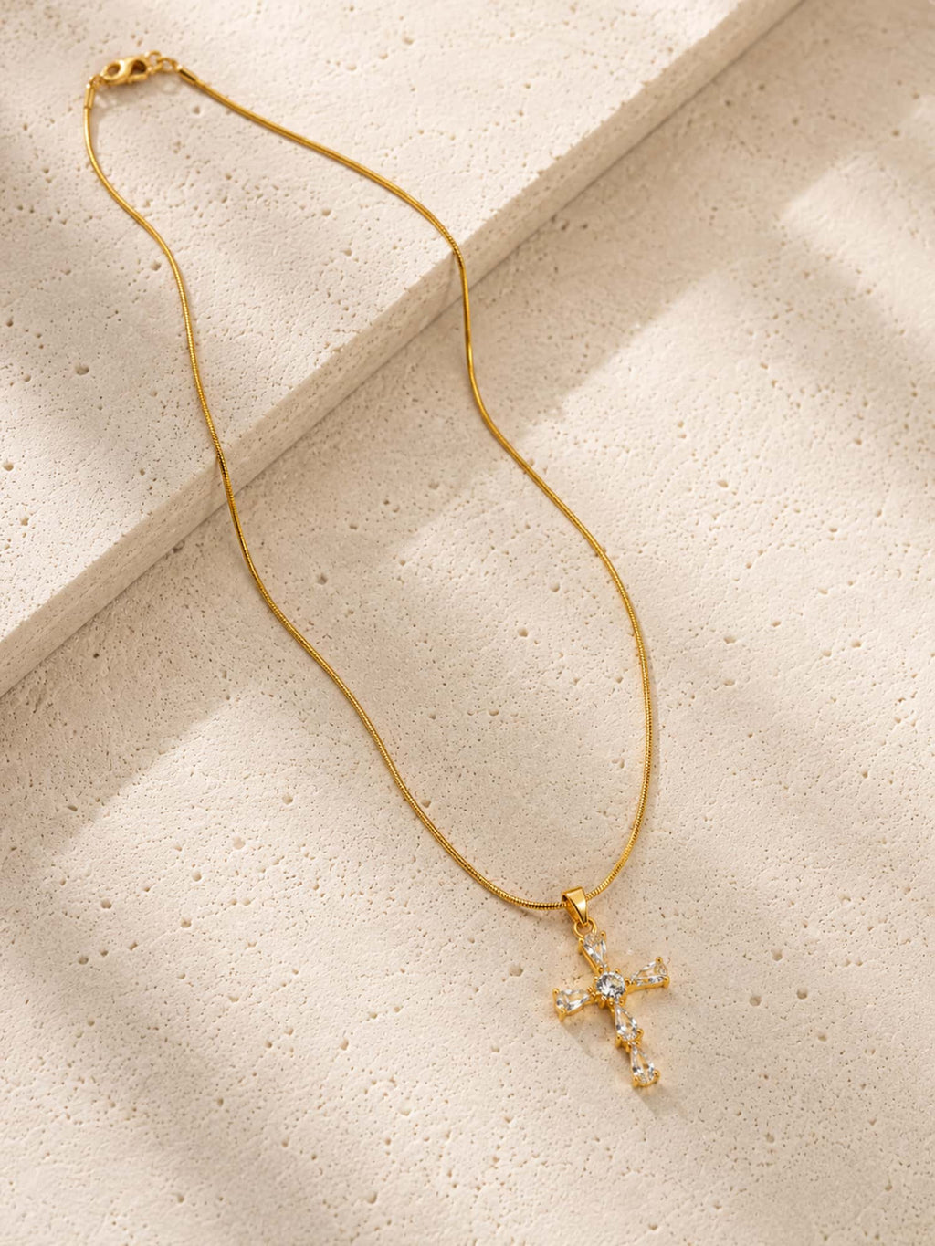 Minimalist CZ cross pendant necklace displayed on a textured stone surface with a gold snake chain.