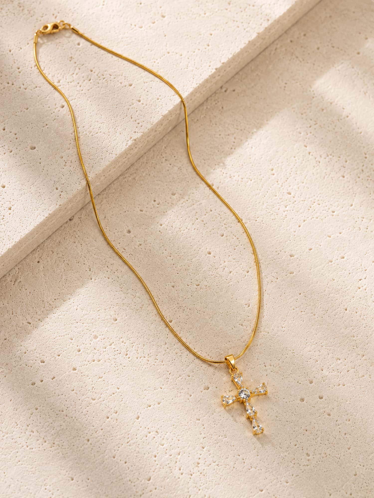 Minimalist CZ cross pendant necklace displayed on a textured stone surface with a gold snake chain.