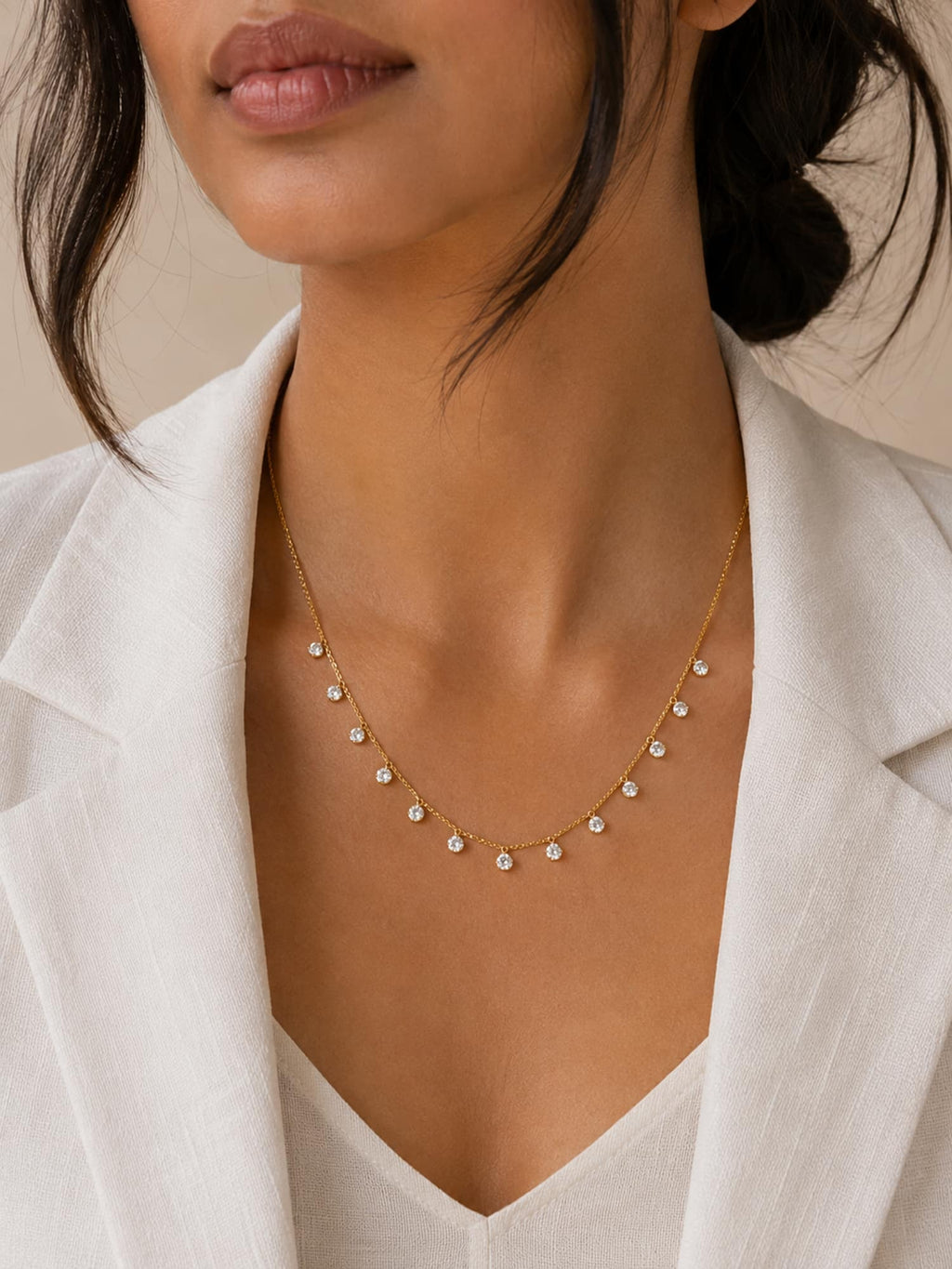 A close-up of a model wearing a delicate gold necklace with small clear gemstone drops, styled with a white linen blazer.