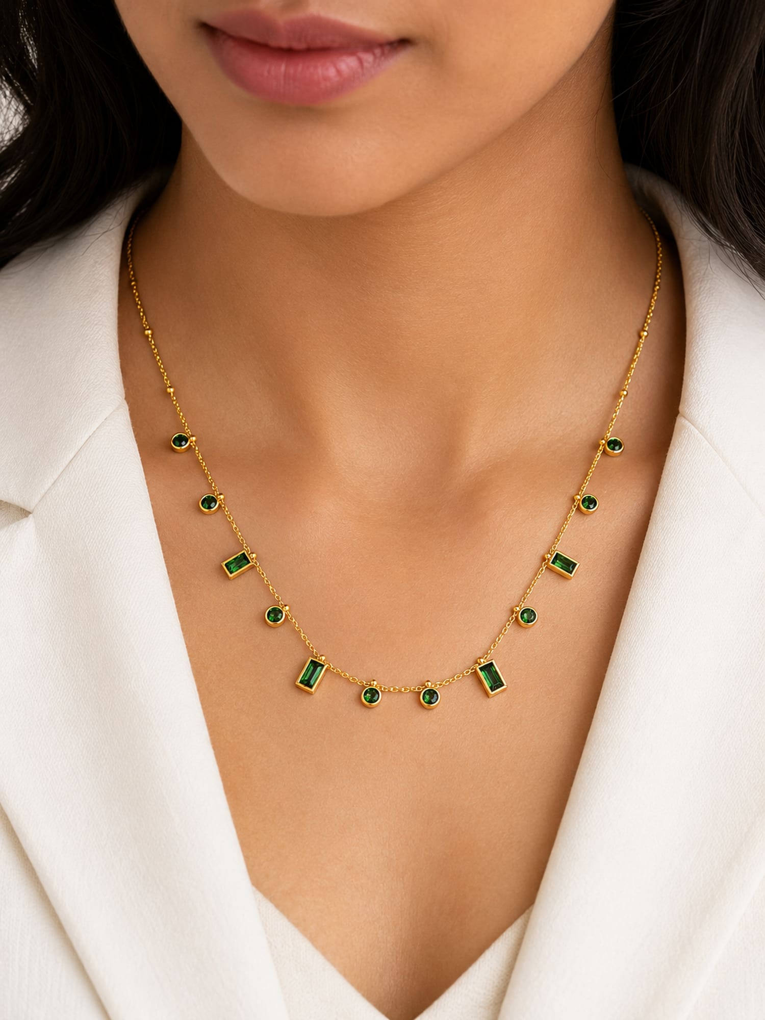A close-up of a model wearing a delicate gold necklace with alternating small round and rectangular green gemstone accents, styled with a white blazer.