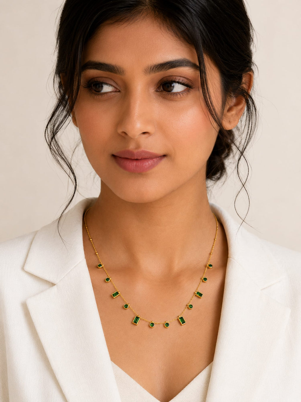 A woman wearing a delicate gold chain necklace with small green rectangular stones, styled with a white blazer against a light beige background.