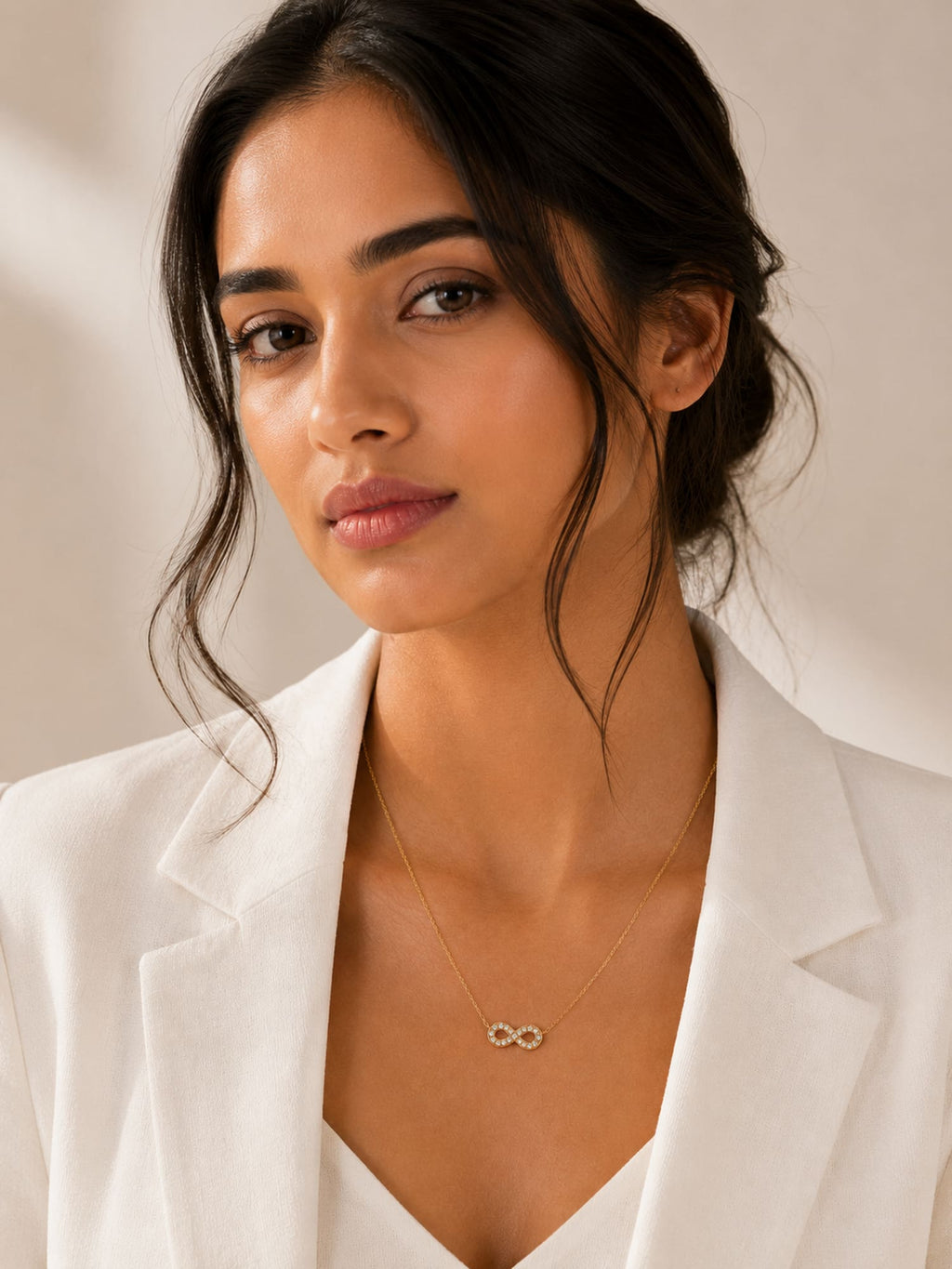 A woman wearing a delicate gold infinity necklace with a small infinity symbol pendant, styled with a white blazer against a soft beige background.