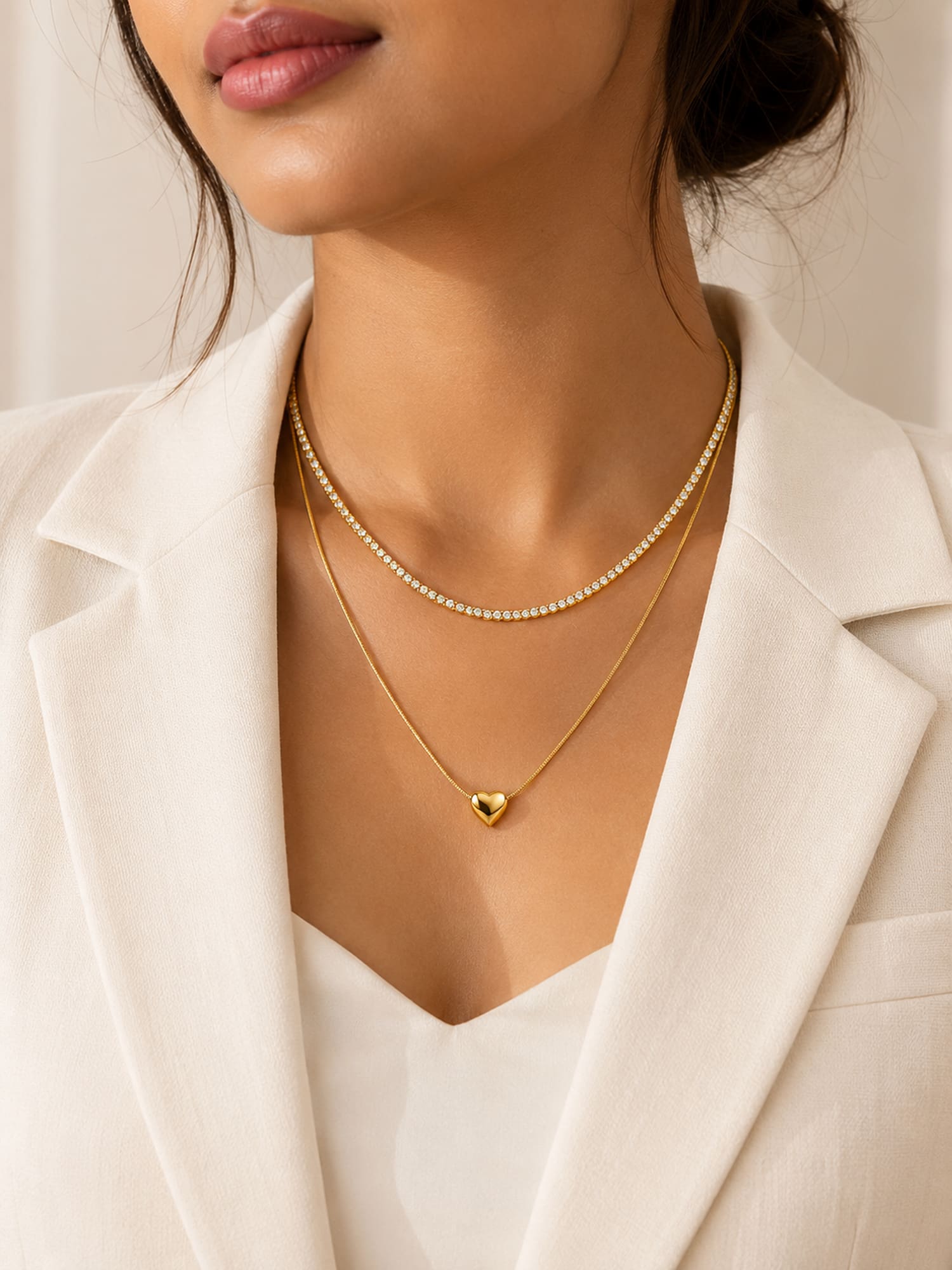 A close-up of a model wearing a double-layered gold necklace with a delicate chain and a small heart pendant, styled with a white blazer.