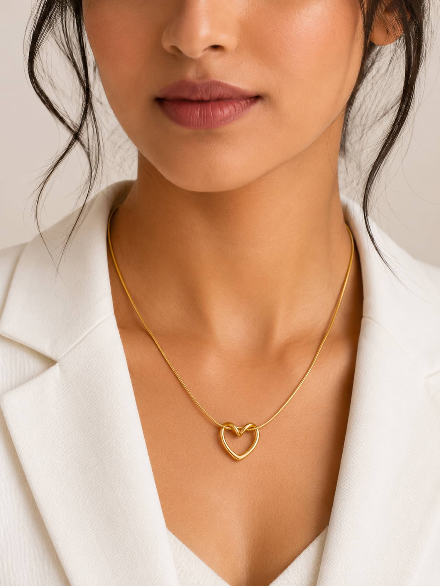 A close-up of a model wearing a delicate gold chain necklace with a small open heart pendant, styled with a white blazer.