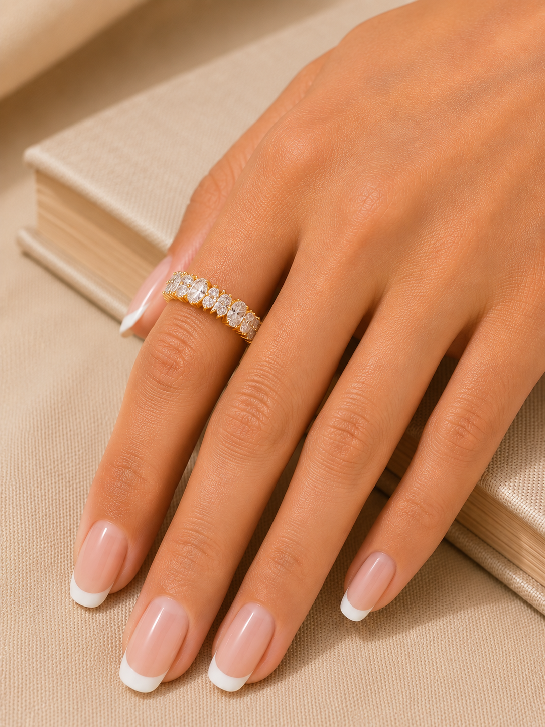 A close-up of a hand with a French manicure wearing a delicate gold ring set with small clear stones, resting on a beige textured surface next to a book.