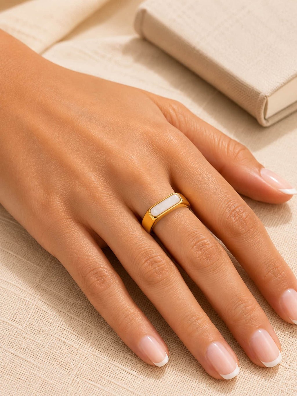 A gold ring with a white inlay, worn on a woman's ring finger, resting on a textured beige fabric surface with a book in the background.