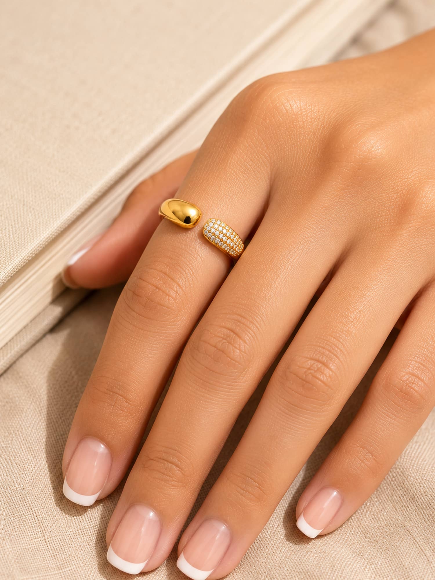 A close-up of a hand wearing two gold rings against a neutral beige textured background. One ring is a smooth polished oval band, and the other is a textured, pavé-set diamond band.