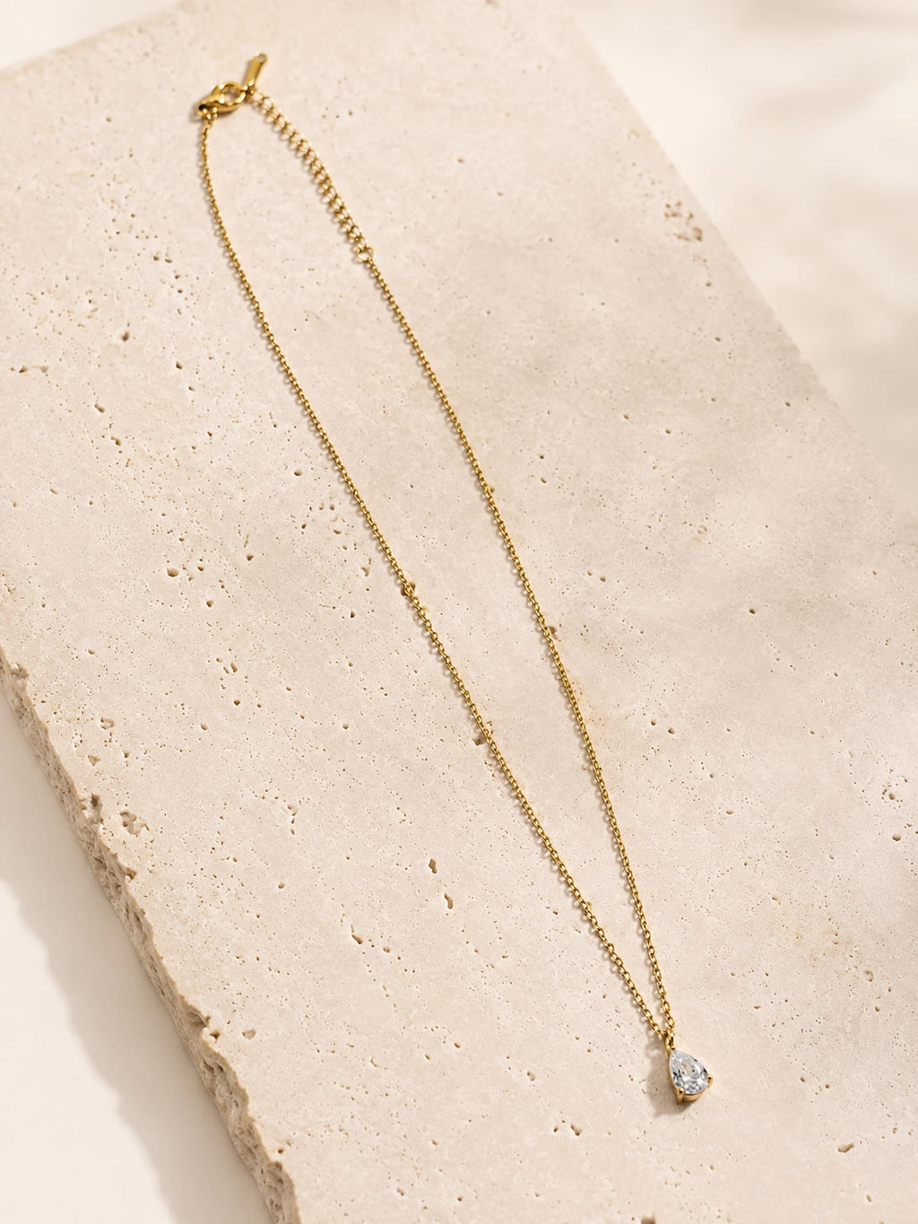 Minimalist pear cut CZ pendant necklace displayed on a textured stone surface in a luxury flat lay.