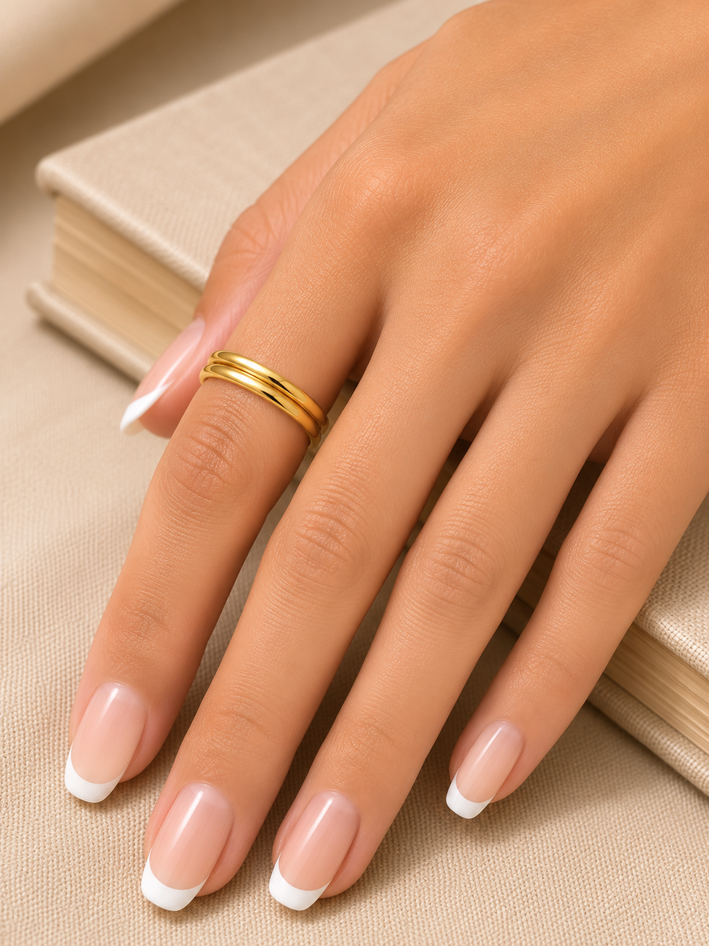 A close-up of a hand with a French manicure wearing a thin double-band gold ring, resting on a beige textured surface next to a book.